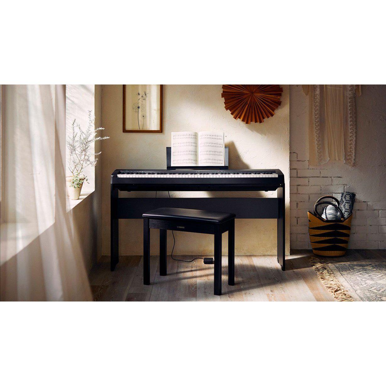 Sort Yamaha digital stagepiano med hvidt nodesæt på stativ, placeret i lyst rum med trægulv og sort klaverbænk foran.
