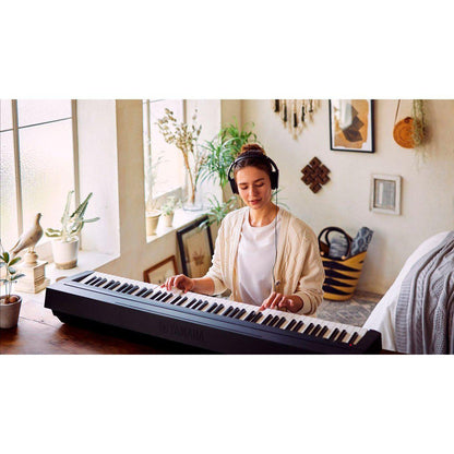Sort Yamaha stagepiano med hvide og sorte tangenter placeret på træbord ved vindue, kvinde med beige cardigan og.