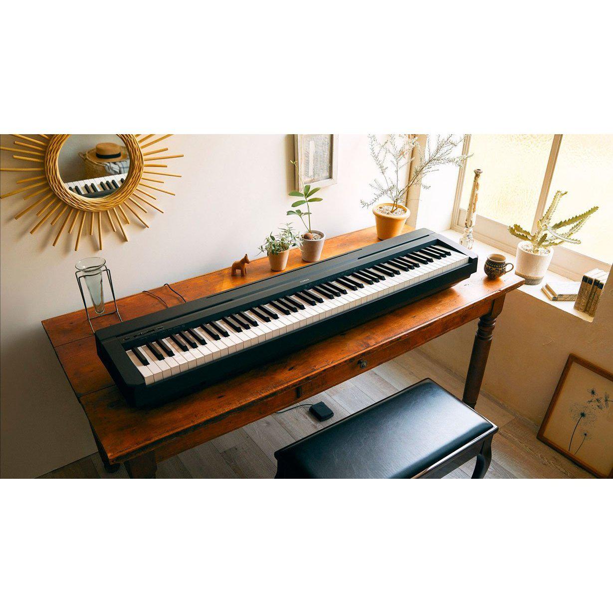 Sort Yamaha stagepiano med 88 tangenter på rustikt træbord ved solbeskinnet vindue med planter og spejl på væggen.