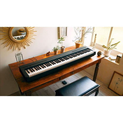 Sort Yamaha stagepiano med 88 tangenter på rustikt træbord ved solbeskinnet vindue med planter og spejl på væggen.